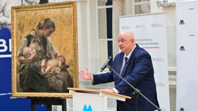 A man at a podium standing next to a painting showing a mother with two children in her lap 