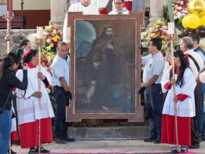 A group of people surrounding a painting of a saint with candles and flowers